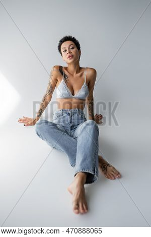 Barefoot Tattooed Woman In Satin Bralette And Jeans Looking At Camera While Sitting On Grey Backgrou