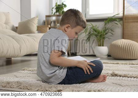 Boy With Poor Posture Reading Book On Beige Carpet In Living Room. Symptom Of Scoliosis