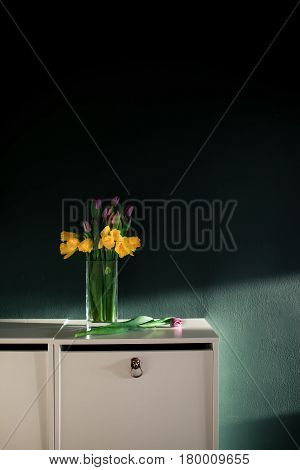 yellow daffodil flowers with pupple tulip blooming in vase with green wall. Dark key. Interior with candle