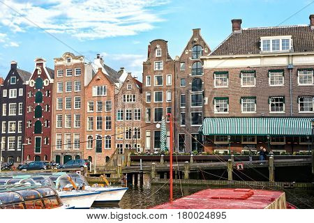 Damrak With Canal Boats And Dancing Houses In Amsterdam