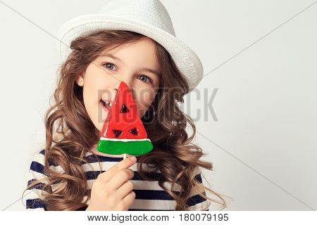 Pretty kid is licking lollipop watermelon shaped. Image on a white studio background.