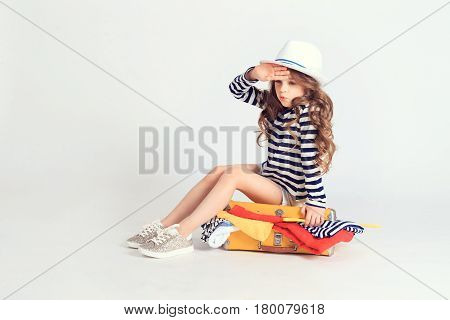 Small tired girl has finished packing her suitcase and sitting on it trying to lock it. A picture is taken at studio and has white background