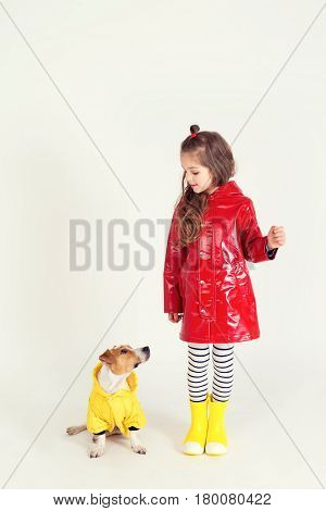 Pretty girl in red raincoat and rubber boots is standing with her dog Jack-Rassel terrier which is wearing yellow raincoat. Kid holding in hand something invisible. hildhood, fashion, imagination, friendship concept