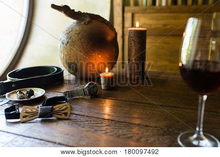 Men's rings in bronze plate, watch and a glass of wine on a wooden table with burning candles. Men's accessorizes, necktie, belt, watch