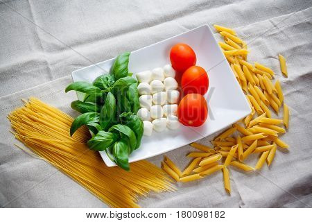 Tomatoes mozzarella raw pasta and green basil leaves on gray tablecloth