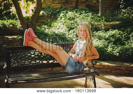 Young girl are sitting on a bench in a park. Blonde female in jeans shorts on the bench with their feet. Rest in park.