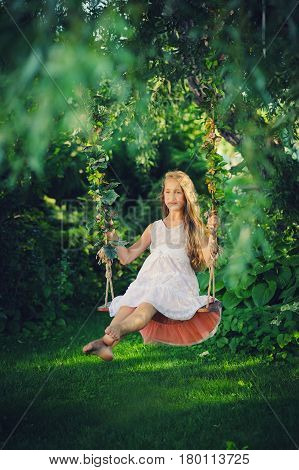Beautiful blond girl on swing on the park with green plants. Girl with long blond hair in dress on swing. Pretty caucasian young girl on the swing with flowers.