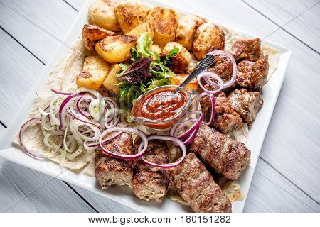 Lyulya kebab with red sauce onion and potatoes on white plate close up with selective focus.