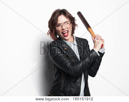 Young trendy girl in black leather coat and glasses posing with batt in hands on white background.