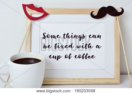 a wooden-framed picture with the text some things can be fixed with a cup of coffee written in it, a red mouth and a mustache attached to sticks and a cup of coffee on a white surface