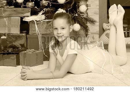 Slender little girl in a white dress lies near the Christmas tree surrounded by boxes with gifts.Black-and-white photo. Retro style.