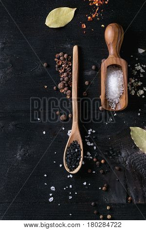 Black himalayan and white sea and fleur de sel salt in wooden spoons with black, chili, allspice pepper and bay leaves over black burnt wood background. Top view with space