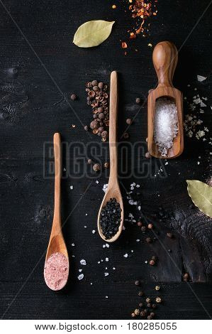 Variety of different colorful salt pink, black himalayan, white sea and fleur de sel in wooden spoons with black, chili, allspice pepper over black burnt wood background. Top view