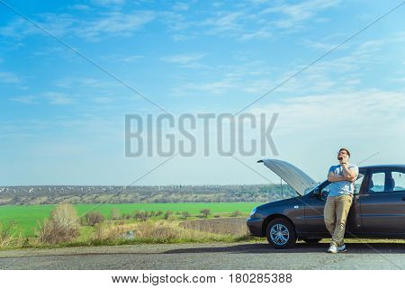 Angry young man waiting a help while sitting near the broken car at the side of road