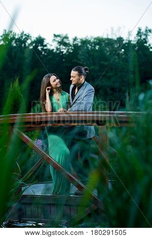 Young people posing on bridge. They are smiling. She in green long dress. He in gray suit.