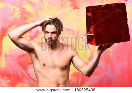 Bearded man short beard. Caucasian sexy macho man with moustache shows six packs and abs on muscle torso holding red package or bag on colorful studio background