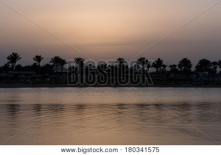 Beautiful sunset or sundown on tropical beach with dark silhouettes of luxury hotel resort place with palms at sea or ocean water in evening dusk on grey sky background. Idyllic summer vacation