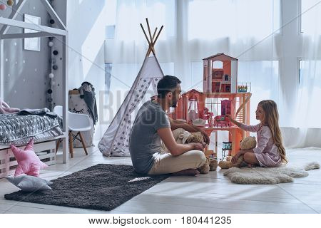 He always understands her. Father and daughter playing with a dollhouse together while sitting on the floor in bedroom