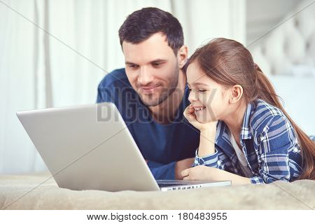 Dad with daughter are staring at the screen of their laptop. Wearing pyjamas they are in bedroom laying on bed.