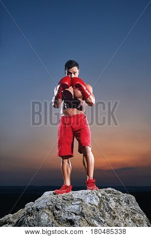 Portrait of a fit young muscular man wearing boxing gloves training outdoors on sunset punching exercising working out preparing competitive motivated determined energetic masculine powerful fighter.