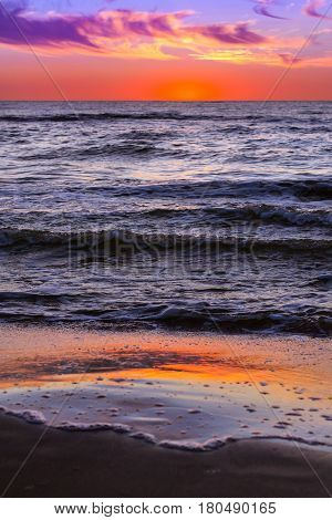 Warm summer night on sandy beach by sea. Sunset at Baltic sea in Lithuanian resort Palanga. Rays of sun shine through the low rare cirrus clouds. Tidal waves wash the sandy beach