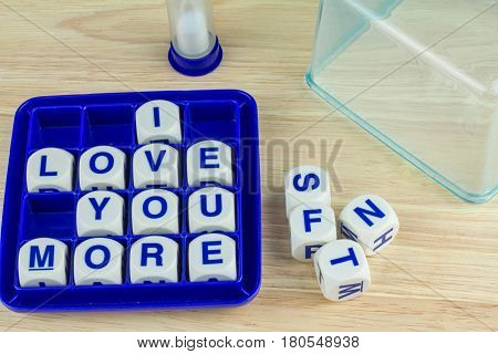 Wording Game with Blue Alphabets on White Cube Blocks say i LOVE YOU MORE in Blue Tray on Wooden Surface Background with Sand Timer and Clear Cover in the back