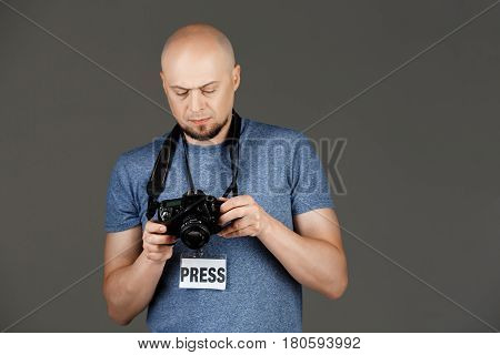 Portrait of handsome middle-aged man in grey shirt with photocamera and press badge taking pictures over dark background. Copy space.