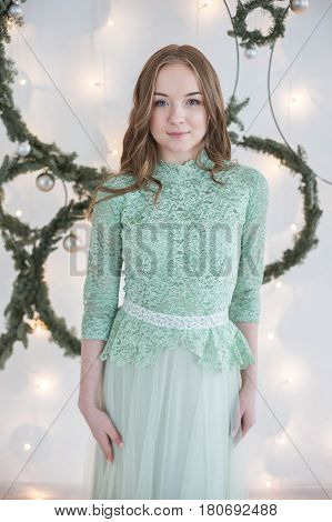 Young beautiful woman with hair of medium length in blue turquoise evening dress in front of a white wall decorated with fairy lights and fir branches poses for the camera.