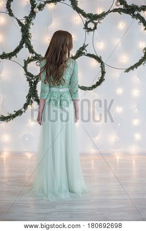 Young beautiful woman with hair of medium length in blue turquoise evening dress in front of a white wall decorated with fairy lights and fir branches poses for the camera.