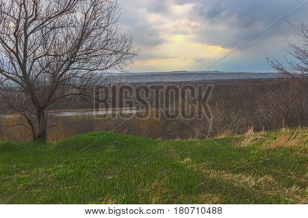 Height on which a last year's grass and also already new green. The river and the wood without foliage is below visible. Further the height and the sky closed by clouds in gleams between which are visible yellow beams of the Sun.