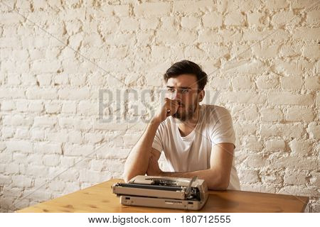 Journalist sit at the table with typewriter and think about new article. Creative crisis concept. Handsome man in white t-shirt hold chin and look away with white brick wall background. Copy space