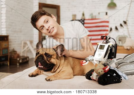 Look and smile. Handsome boy keeping smile on his face lying behind his pet while scratching his ear