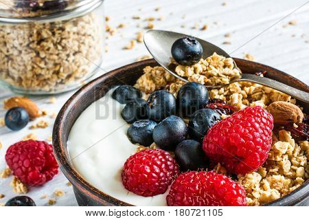 bowl of oat granola with yogurt fresh raspberries blueberries and nuts with cereals and spoon in a jar for healthy breakfast. close up