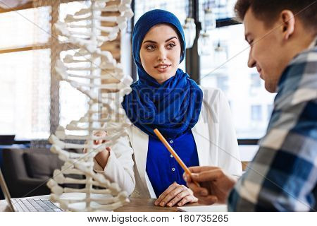 Interesting process. Pleasant smilign muslim girl sitting at the table with her groupmate and examinign DNA model while studying genetics