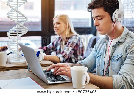Concentrate on work. Pleasant involved student using laptop and sitting on the cafe while his friends learnign genetics in the background