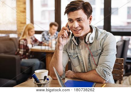 Nice to hear you. Positive delighted smiling young man sitting at the table and talking on smart phone while students learnign in the background