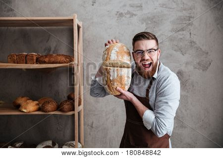 Picture of screaming young man baker standing at bakery holding bread. Looking at camera.