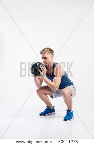 Handsome hipster fitness man in blue tank top shirt and gray shorts holding medicene ball, doing squat. Studio shot on gray background.