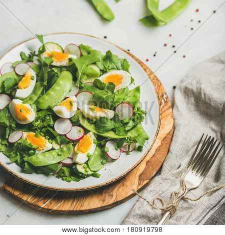 Healthy spring green salad with radish, boiled egg, arugula, green pea and mint in white plate on olive tree wood board over grey background, selective focus, square crop