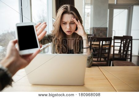 Serious young woman discarding the phone while using laptop in cafe. Phone with blank screen
