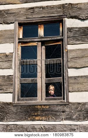 Small and lonely caucasian baby girl or toddler looking out of old wooden cabin window to suggest poverty or recession