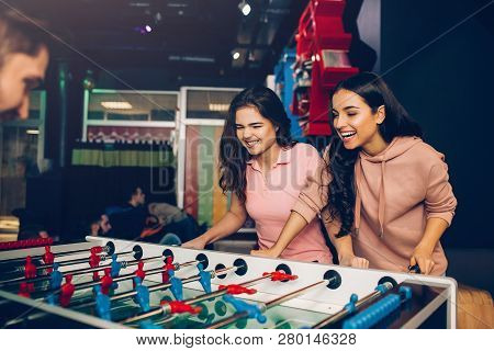 Playful Excited Young Women Playing Table Soccer With Guy In Room. Models Loook At Game And Control 