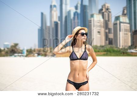 Portrait Of Young Woman In Bikini, Sunglasses And Straw Hat Standing At The Beach With Skycrappers O