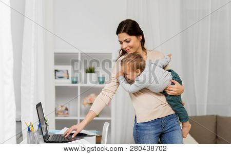 motherhood, multi-tasking, family and technology concept - happy mother with baby, laptop computer and papers working at home