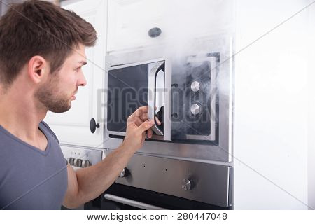 Astonished Man Standing In Front Of Burning Oven With Smoke Around Kitchen
