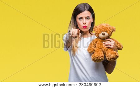 Young beautiful woman holding teddy bear plush over isolated background pointing with finger to the camera and to you, hand sign, positive and confident gesture from the front