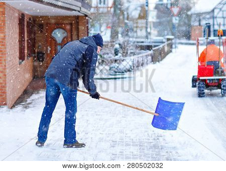 Man With Snow Shovel Cleans Sidewalks In Winter During Snowfall. Winter Time In Europe. Young Man In
