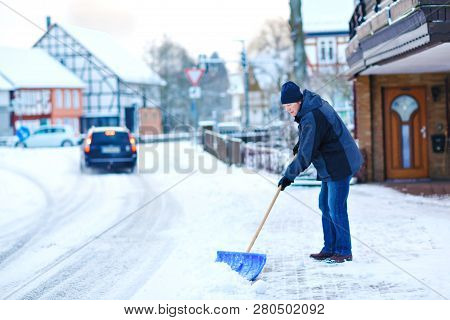 Man With Snow Shovel Cleans Sidewalks In Winter During Snowfall. Winter Time In Europe. Young Man In