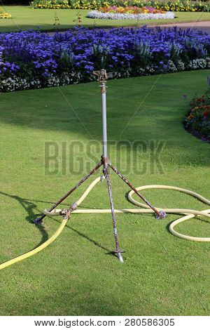Water Sprinkler With Attached Yellow Hosepipe Standing On A Mown Grass Lawn With Summer Flower Beds 