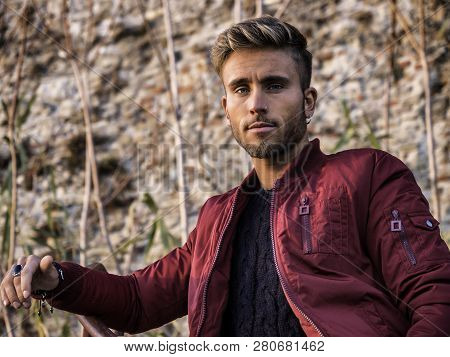 Handsome Young Man Outdoor In Winter Fashion, Wearing Red Jacket And Woolen Scarf In City Park, Look